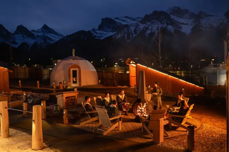 People gather around a fire pit in the Fire Gardens, with yurts and mountains in the background.