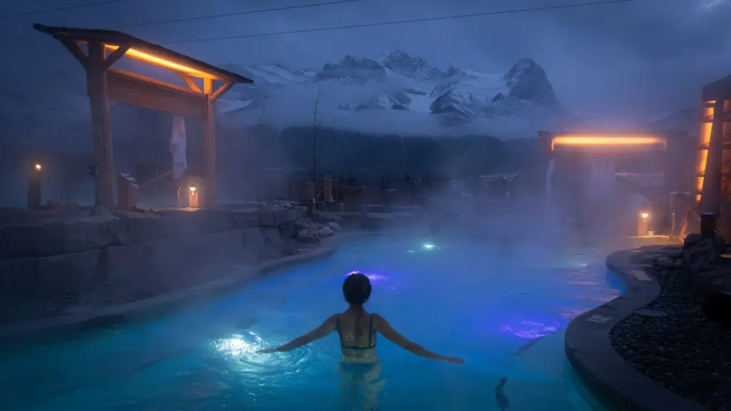 Woman relaxing in a steamy outdoor hot spring at night with snowy mountains
