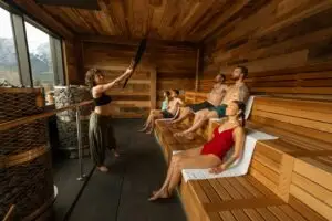 People relaxing in a wooden sauna with a woman adding water to hot stones.